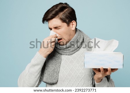 Young ill sick man wear gray sweater scarf sneezing hold box of paper napkins sneeze isolated on plain blue background studio. Healthy lifestyle disease virus treatment cold season recovery concept