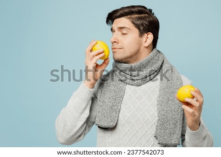 Young ill sick man wearing gray sweater scarf hold sniff citrus lemon fruit isolated on plain blue background studio portrait. Healthy lifestyle disease virus treatment cold season recovery concept