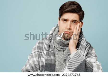 Young ill sick man wrapped in gray plaid keep thermometer in mouth hold cheek isolated on plain blue background studio portrait. Healthy lifestyle disease virus treatment cold season recovery concept