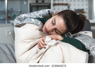 Young ill woman wrapped in blanker blowing her nose in to the toilet paper. Sick female sitting at home on sofa try to warm up suffering from common cold or flu, feeling exhausted and tired.
