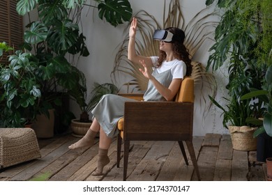 Young impressed woman wearing VR helmet sitting in armchair at greenhouse, monstera deliciosa plant on background. Hispanic female experiencing virtual reality while resting in beautiful indoor garden - Powered by Shutterstock