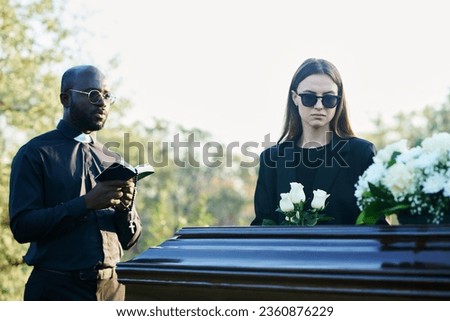 Young inconsolable widow in mourning attire and sunglasses holding bunch of white roses while standing by coffin next to priest with Bible