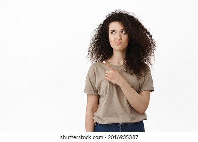 Young Indecisive Woman Pointing And Looking At Upper Left Corner, Making Perplexed Face Expression, White Background