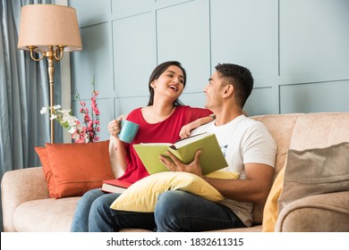 Young Indian Asian Couple Reading Books While Sitting On Sofa At Home, Modern Living Space