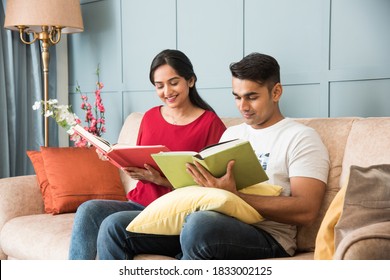 Young Indian Asian Couple Reading Books While Sitting On Sofa At Home, Modern Living Space