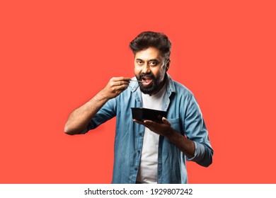 Young Indian Asian Man Eating Take Away Food In The Box Using Spoon Or Chopsticks, Standing Isolated Against Red Background