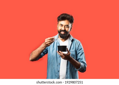 Young Indian Asian Man Eating Take Away Food In The Box Using Spoon Or Chopsticks, Standing Isolated Against Red Background