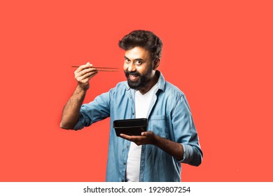 Young Indian Asian Man Eating Take Away Food In The Box Using Spoon Or Chopsticks, Standing Isolated Against Red Background