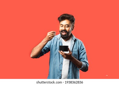 Young Indian Asian Man Eating Take Away Food In The Box Using Spoon Or Chopsticks, Standing Isolated Against Red Background