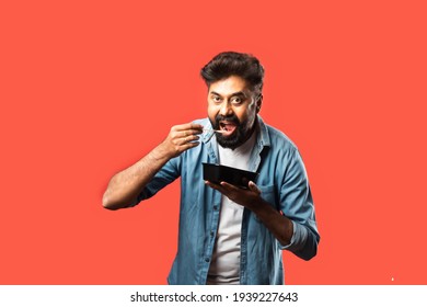 Young Indian Asian Man Eating Take Away Food In The Box Using Spoon Or Chopsticks, Standing Isolated Against Red Background