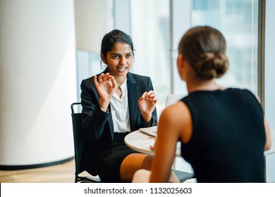 A young Indian Asian woman has a business meeting (interview) with a Caucasian white woman in an office during the day. They are both young, attractive and professionally dressed. 