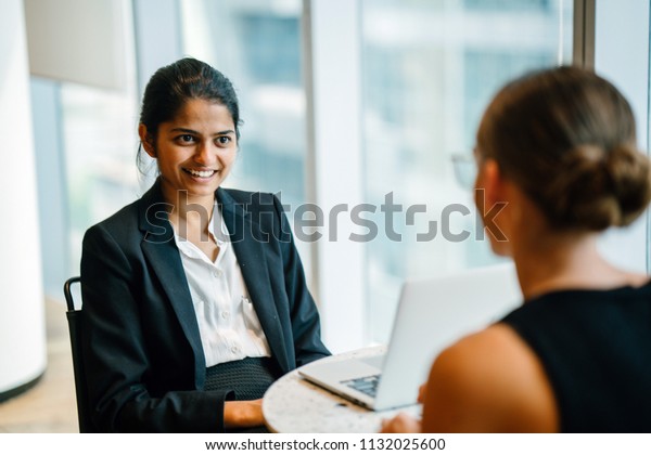 A young\
Indian Asian woman has a business meeting (interview) with a\
Caucasian white woman in an office during the day. They are both\
young, attractive and professionally dressed.\

