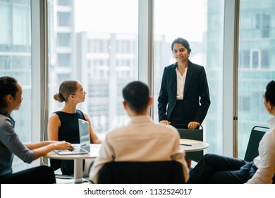 A Young Indian Asian Woman Stands Up In Front Of Her Diverse Team And Is Leading A Meeting, Training Or Presentation In Their Office During The Daytime. They Are An Ethnically Diverse Team.