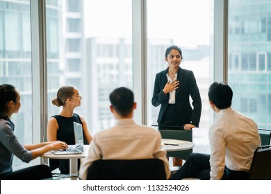 A Young Indian Asian Woman Stands Up In Front Of Her Diverse Team And Is Leading A Meeting, Training Or Presentation In Their Office During The Daytime. They Are An Ethnically Diverse Team.