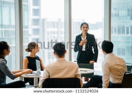 A young Indian Asian woman stands up in front of her diverse team and is leading a meeting, training or presentation in their office during the daytime. They are an ethnically diverse team.