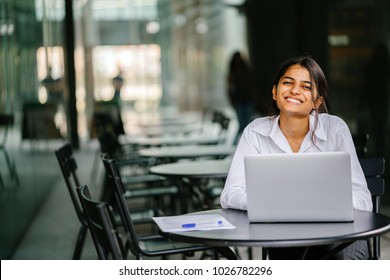 A Young Indian Asian Woman Is Typing And Working In A Laptop In The Day (applying For A Job, Etc). She Is Wearing A Professional, Crisp White Shirt And She Is Smiling As She Types On Her Laptop. 