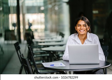 A young Indian Asian woman is typing and working in a laptop in the day (applying for a job, etc). She is wearing a professional, crisp white shirt and she is smiling as she types on her laptop. 