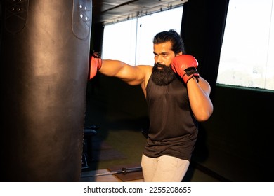 Young indian beard man wearing boxing gloves and hooded sweatshirt punching bag in gym, Fitness and healthcare. - Powered by Shutterstock