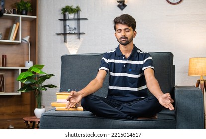 Young Indian Boy Meditating With Eyes Closed Before Reading While Sitting On Sofa At Home - Concept Of Stress Relief, Knowledge And Relaxation.