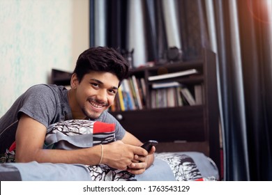 Young Indian boy smiling into the camera, while using his phone lying on his bed.