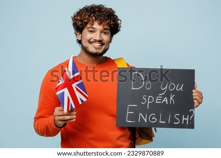 Young Indian boy student wear casual clothes bag hold British flag card with text do you speak english travel abroad to study isolated on plain blue background High school university college concept