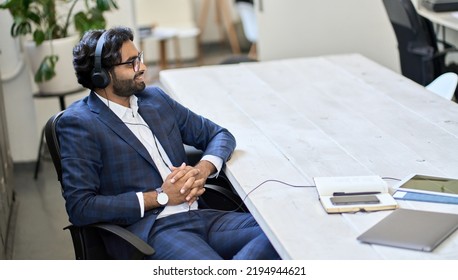 Young Indian Business Man Professional Employee Or Executive Manager Wearing Headphones Sitting At Workplace Listening To Music Or Business Podcast, Having Remote Virtual Call Working In Office.