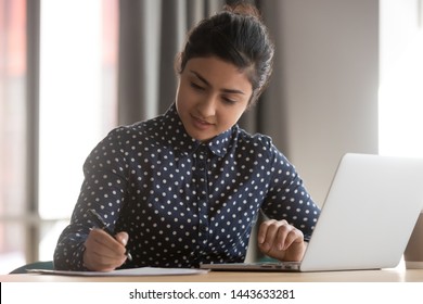 Young indian business woman student working studying online with laptop making notes sit at office desk, focused hindu female professional preparing report doing paperwork writing essay at workplace