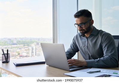 Young Indian Businessman Ceo Executive Sitting At Desk Using Laptop Near Panoramic Window In Contemporary Office. Serious Financial Analyst Working With Online Data. Business Technology Concept.