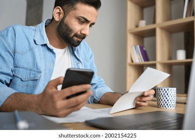Young indian businessman holding phone reading bank receipt calculating taxes  ethnic man using smartphone mobile application checking bill document  managing money finances  loan expenses 
