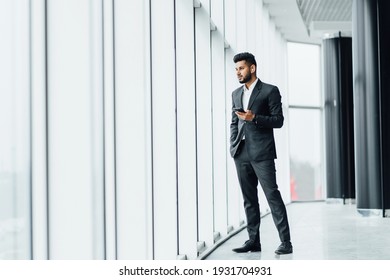 A young Indian businessman in a modern office building, he is standing by the window and talking on the phone, a business meeting, wearing a black suit.