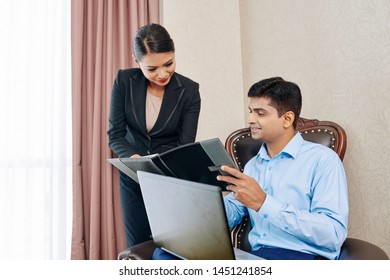 Young Indian Businessman Sitting On Chair With Laptop Computer And Reading The Rule Of The Hotel In Folder Giving By The Asian Manager