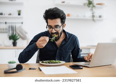 Young indian businessman tasting fresh greens in vegan meal while sitting at office desk in bright apartment. Self-employed worker enjoying nutritious food during lunch hour while telecommuting. - Powered by Shutterstock