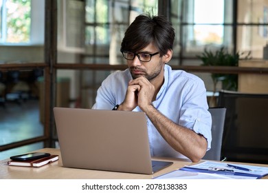 Young Indian Businessman Using Computer Working Online Watching Business Training Webinar In Office. Ethnic Guy Employee Thinking On Project Analysis Looking At Laptop At Workplace.