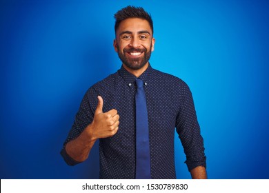 Young indian businessman wearing elegant shirt and tie standing over isolated blue background doing happy thumbs up gesture with hand. Approving expression looking at the camera showing success. - Shutterstock ID 1530789830