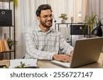 Young Indian businessman wearing headset, freelance worker, call center or support service operator helpline, talking with client or colleague communication support. Guy sitting at home office desk.