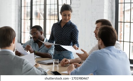 Young Indian businesswoman lead meeting with diverse colleagues share handouts at briefing, confident female biracial team leader head team discussion in office, consider paperwork with coworkers - Shutterstock ID 1701980467