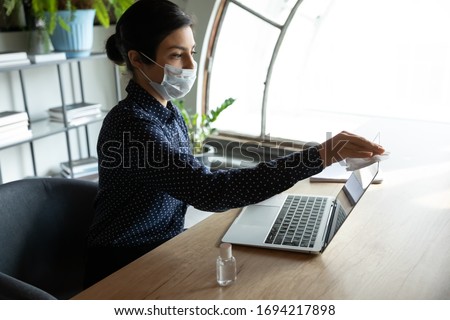 Young indian businesswoman using antiseptic sanitizer, cleaning computer screen monitor before starting working in office. Female employee in breath protective facemask disinfecting workplace.