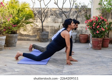 A young Indian couple dressed in sportswear, perform synchronized yoga together, practicing on their terrace surrounded by flower pots, against the clear blue sky on a sunny morning in New Delhi.