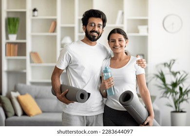 Young indian couple holding yoga mats at home and smiling at camera, eastern spouses going to gym together, happy man and woman in sportswear posing with fitness mats and water bottle in hands - Powered by Shutterstock