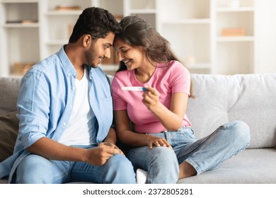 Young indian couple radiating joy while comfortably seating on sofa, sharing tender moment of happiness, holding positive pregnancy test and touching their foreheads - Powered by Shutterstock