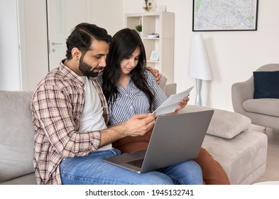Young indian couple reading paper bills paying loan bank debt online together on computer, calculating taxes, income, making payments, planning family budget money finances using laptop at home. Foto Stock