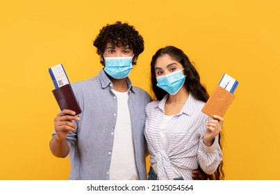 Young Indian Couple Ready To Travel Abroad During COVID-19 Pandemic, Wearing Protective Face Masks, Holding Passports And Flight Tickets, Yellow Studio Background