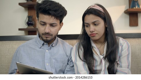 A Young Indian Couple With Serious Facial Expression Looking At The Tablet While Sitting On The Sofa