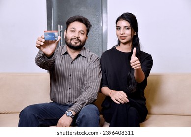 Young indian couple showing credit card and giving expression while sitting on sofa - Shutterstock ID 2396408311