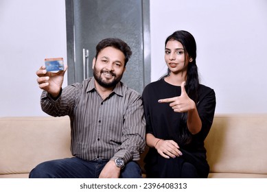 Young indian couple showing credit card and giving expression while sitting on sofa - Shutterstock ID 2396408313