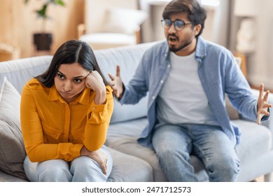 A young Indian couple is sitting on a couch in their living room engaged in a heated argument. The woman appears upset and is looking away, while the man is gesturing passionately with his hands. - Powered by Shutterstock