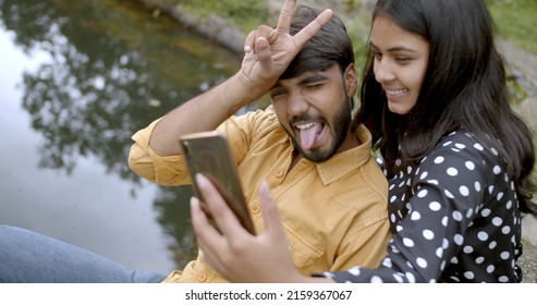 A Young Indian Couple Taking A Wacky Selfie By A Pond At A Park