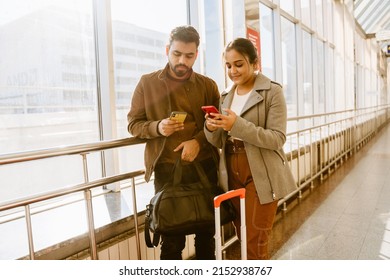 Young Indian Couple Using Cellphones While Standing In Airport Indoors