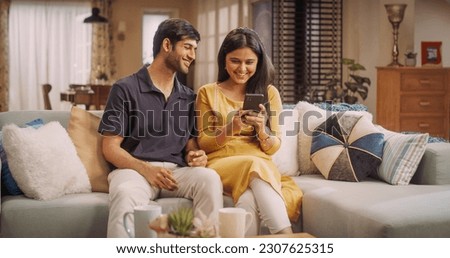 Young Indian Couple Using Internet On Smartphone, Sitting On The Sofa at Home, they Tease and Joke around. Fun Boyfriend and Girlfriend Choosing Products, Doing Online Shopping.