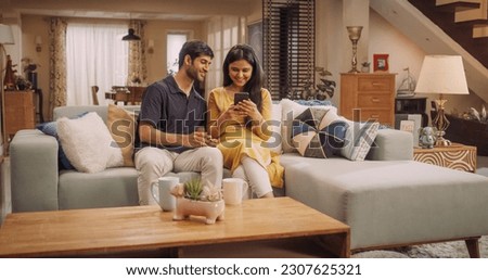 Young Indian Couple Using Internet On Smartphone, Sitting On The Sofa at Home, they Tease and Joke around. Playful Boyfriend and Girlfriend Choosing Products, Doing Online Shopping.